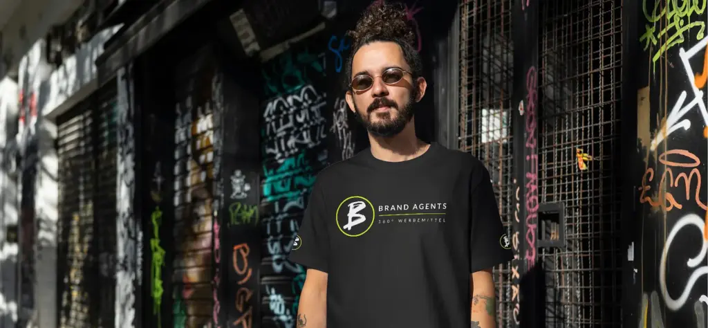 Mann trägt ein schwarzes T-Shirt mit dem Logo von Brand Agents vor einer graffiti-bedeckten Wand.