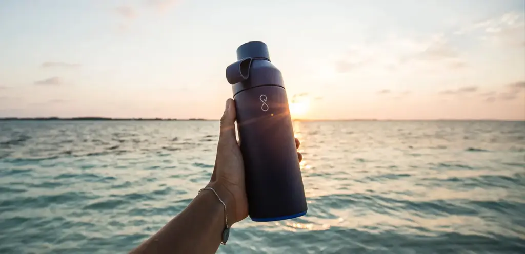 Schwarze Wasserflasche in der Hand vor einem Sonnenuntergang über dem Wasser.
