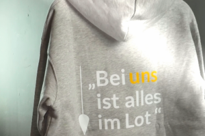 Grauer Hoodie mit dem Text 'Bei uns ist alles im Lot' auf der Rückseite in gelber und grauer Schrift.