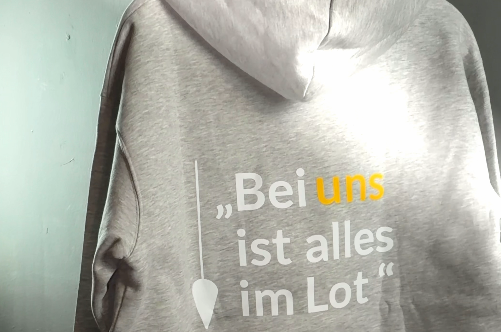 Grauer Hoodie mit dem Text 'Bei uns ist alles im Lot' auf der Rückseite in gelber und grauer Schrift.