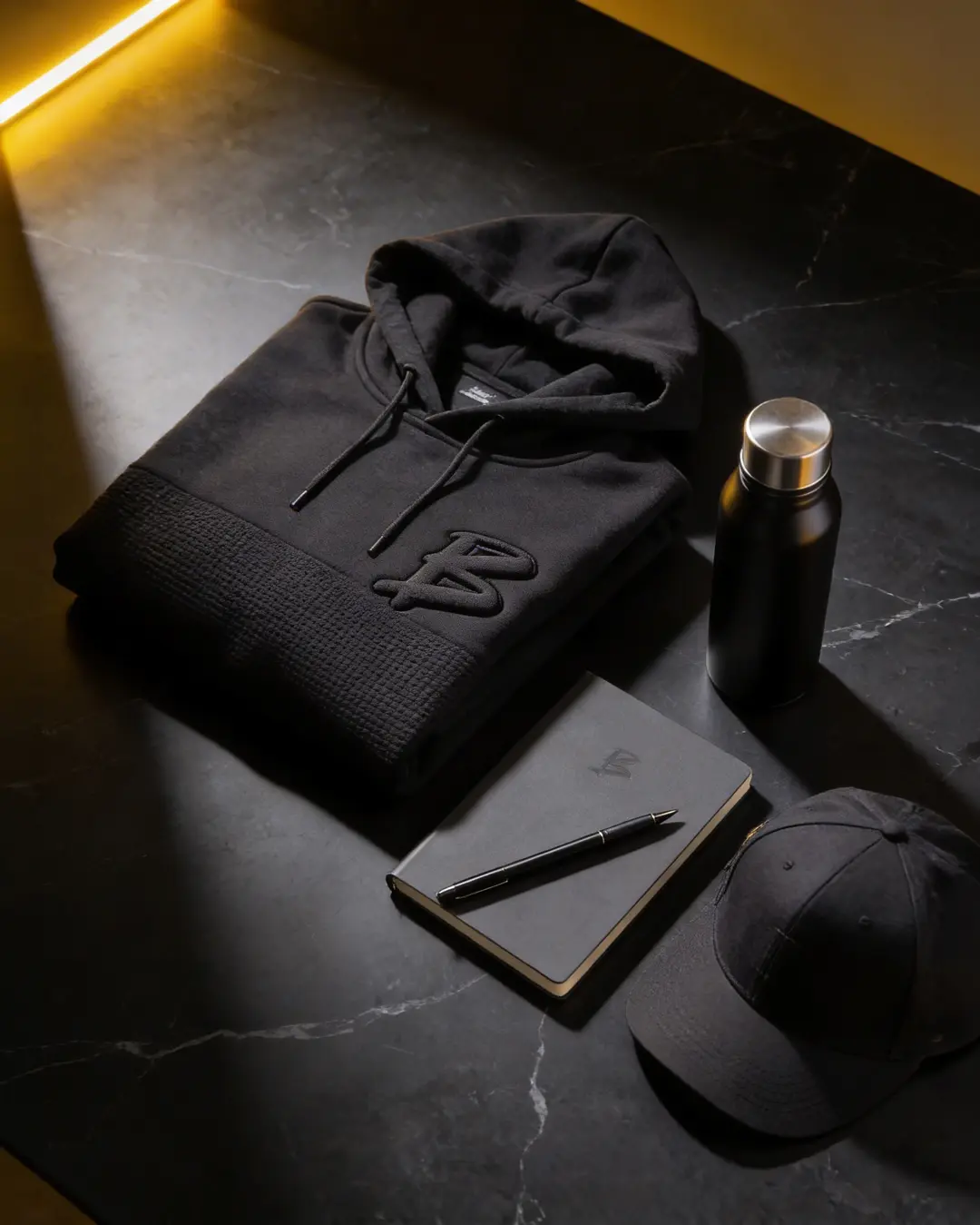 Schwarzer Kapuzenpullover mit gesticktem Logo, eine schwarze Wasserflasche, ein Notizbuch und eine schwarze Kappe auf einem Tisch.