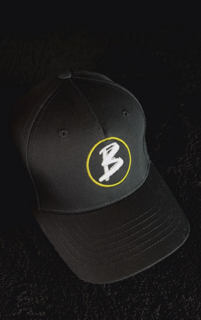 Schwarze Baseballkappe mit gelbem Kreis und weißem Schriftzug 'B' auf der Vorderseite.