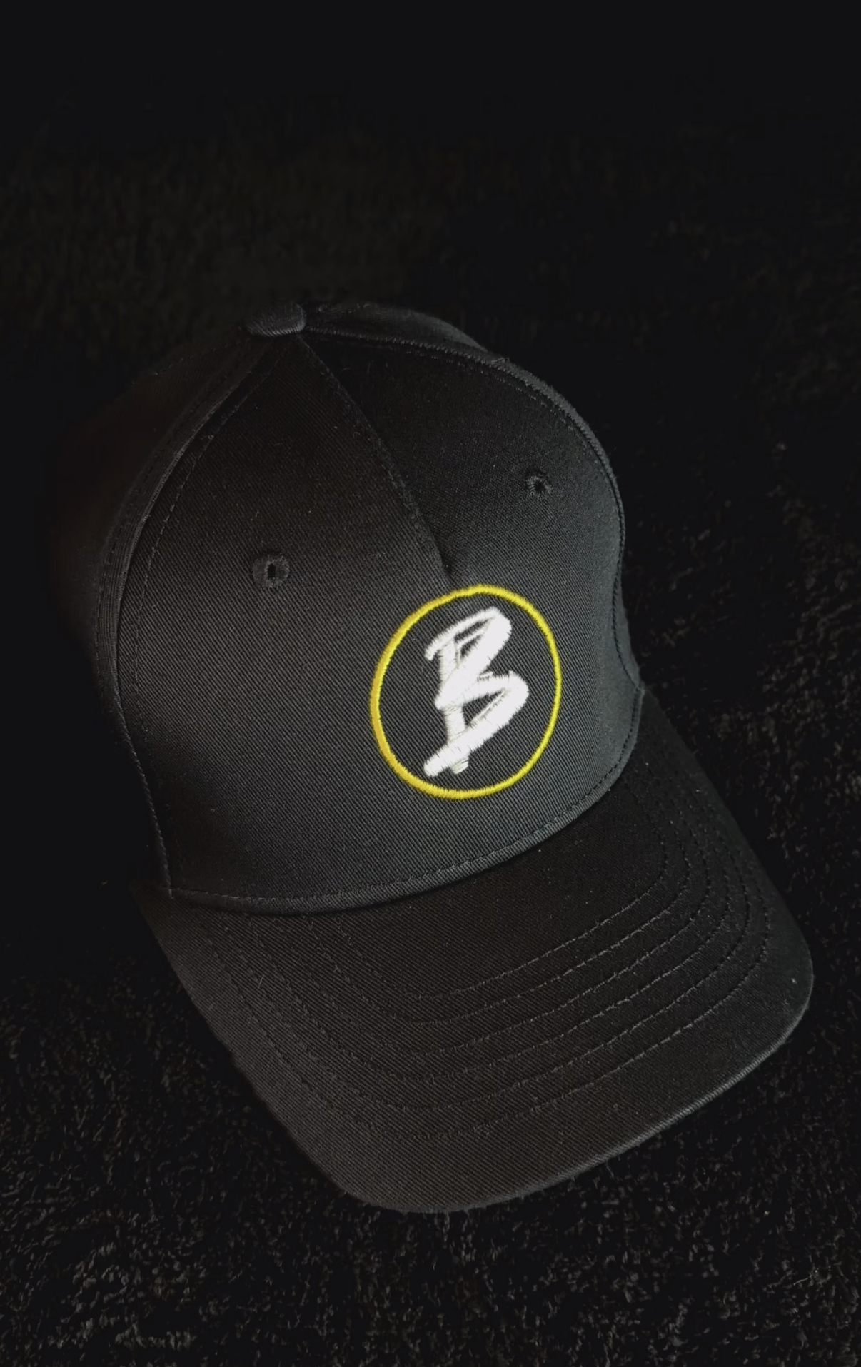 Schwarze Baseballkappe mit gelbem Kreis und weißem Schriftzug 'B' auf der Vorderseite.