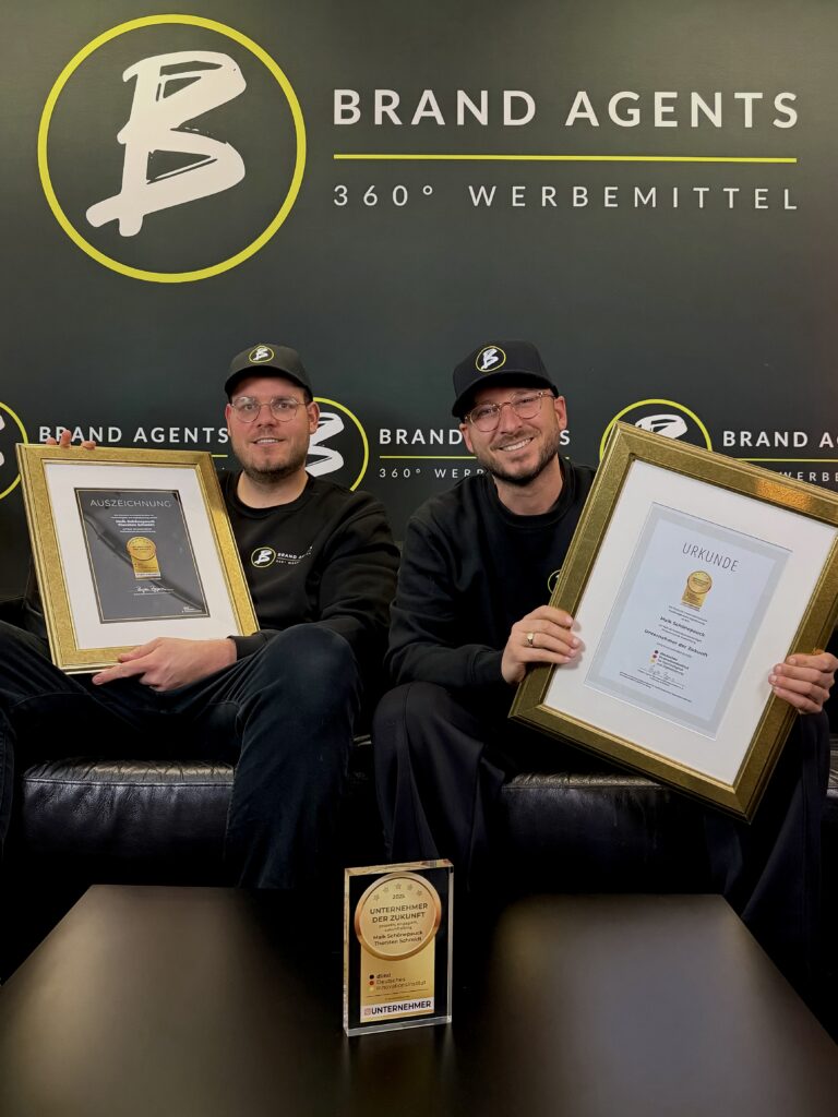 Zwei Männer in Schwarz tragen Brillen und Caps mit Brand Agents Logo, halten gerahmte Urkunden; Hintergrund mit Brand Agents Branding und gelber Akzentlinie, Kleiner Pokal auf dem Tisch