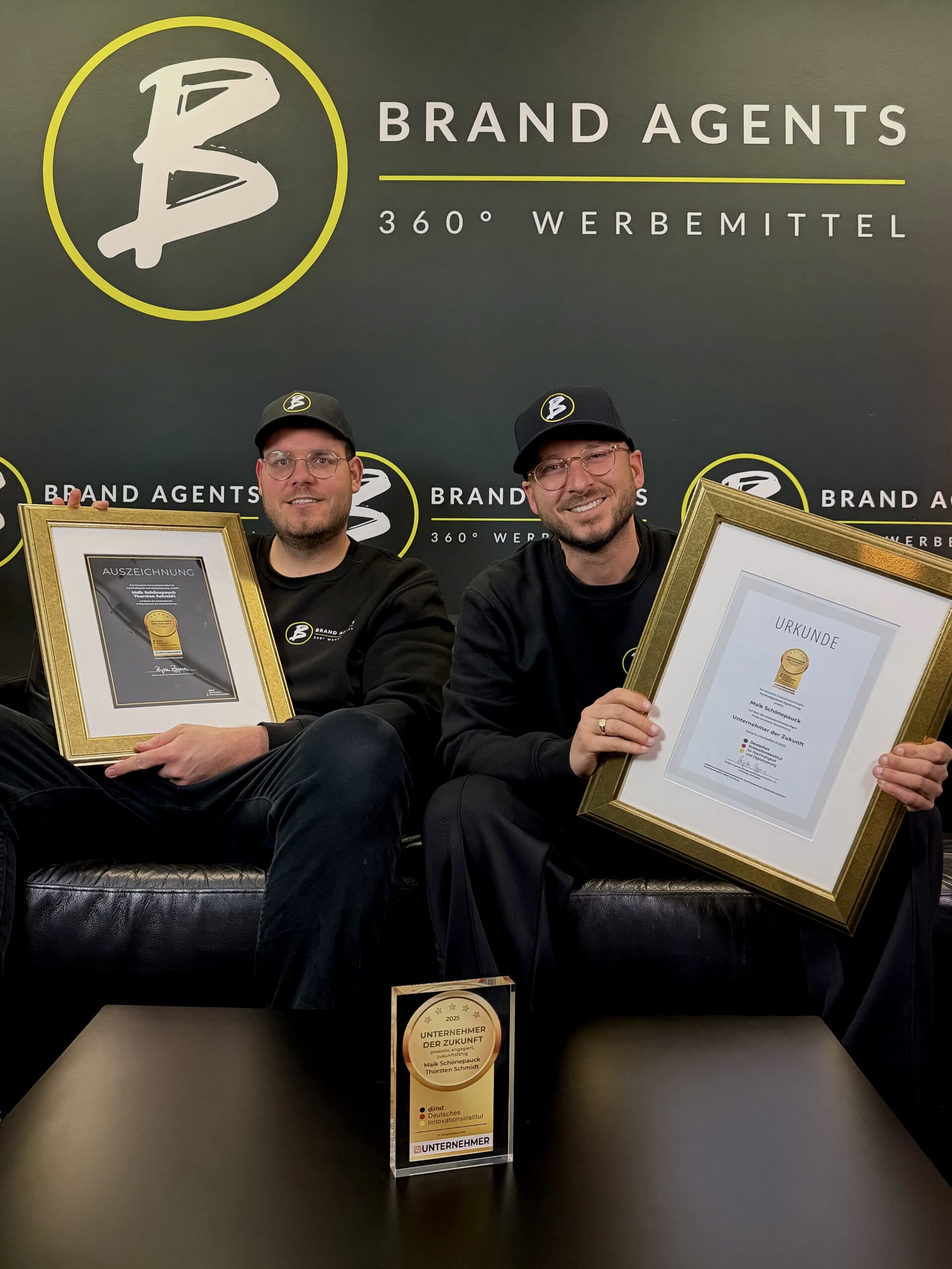 Zwei Männer in Schwarz tragen Brillen und Caps mit Brand Agents Logo, halten gerahmte Urkunden; Hintergrund mit Brand Agents Branding und gelber Akzentlinie, Kleiner Pokal auf dem Tisch