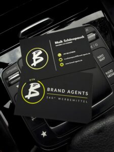 Brand Agents Visitenkarten mit gelbem Logo auf schwarzem Hintergrund, Maik Sch&ouml;nepauck genanntes Branding, kompakte Karten im Autoinnenraum.