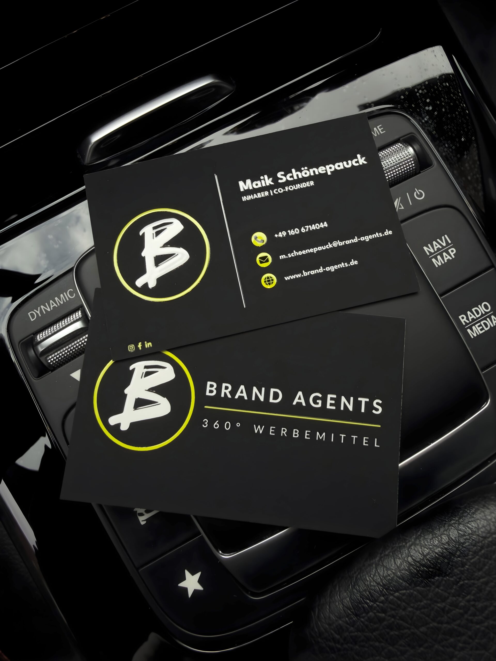 Brand Agents Visitenkarten mit gelbem Logo auf schwarzem Hintergrund, Maik Schönepauck genanntes Branding, kompakte Karten im Autoinnenraum.