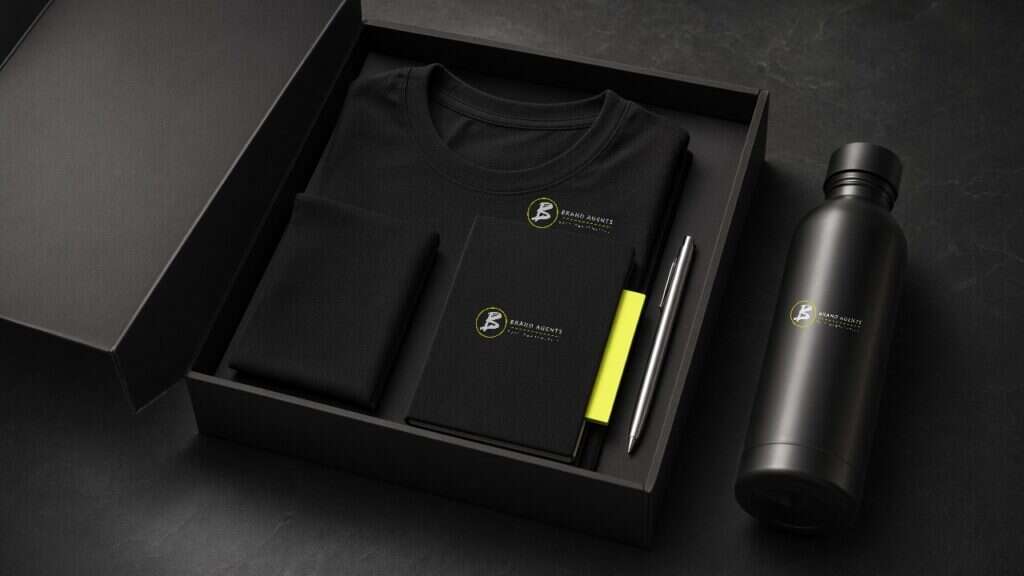 Schwarzes Willkommenspaket mit Oberteil, Notizbuch und Trinkflasche mit Brand-Agents-Logo sowie einem silberfarbenen Stift.